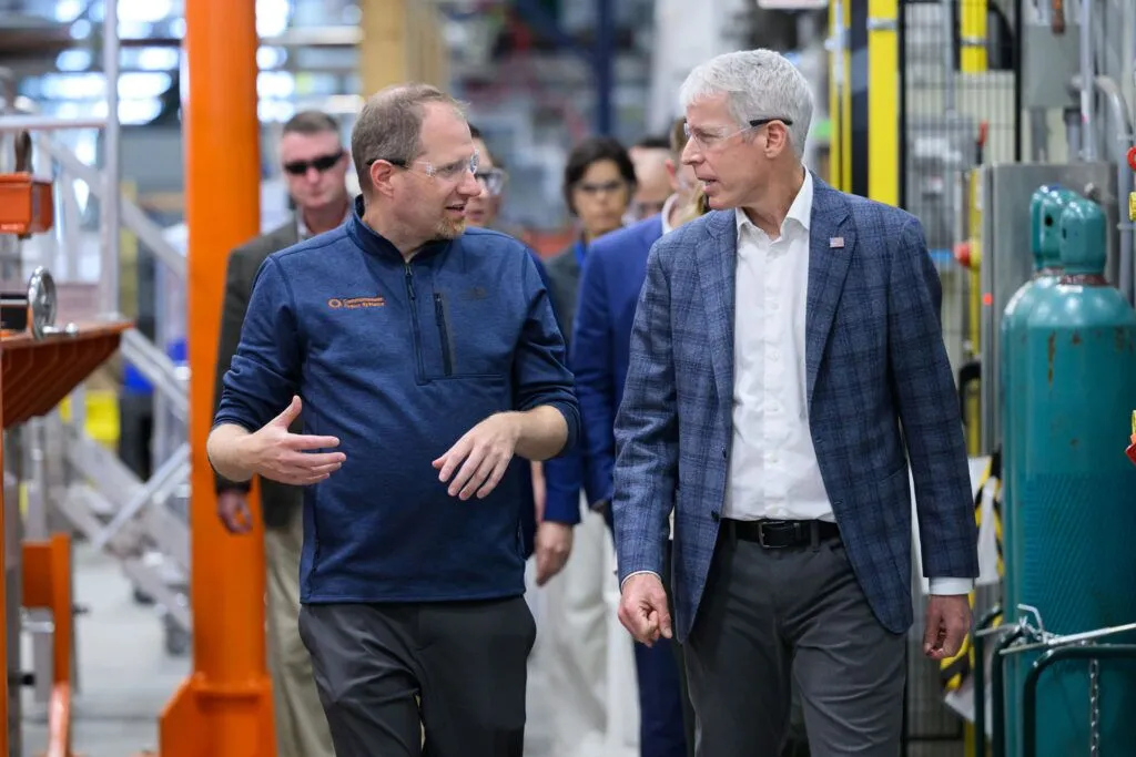 Two men wearing protective eyewear walk through a factory floor, orange metal structures on one side and green tanks of gas on the other.