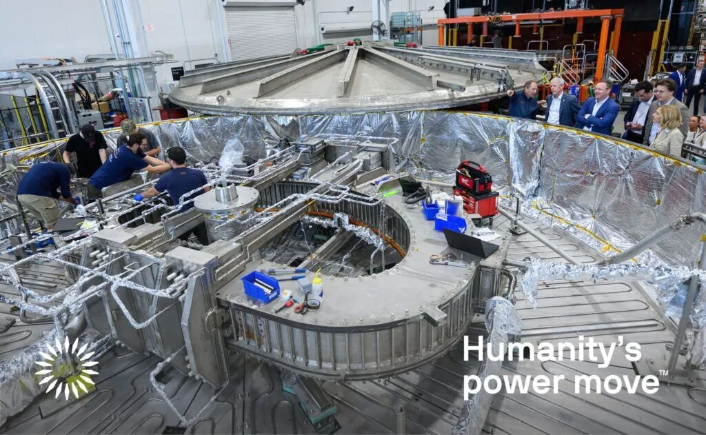 A group of people discuss a large, wide silvery cylindrical test chamber that houses a D-shaped metal structure in its center. Five workers inside the chamber make adjustments. A number of tools and computers nearby support the operation 