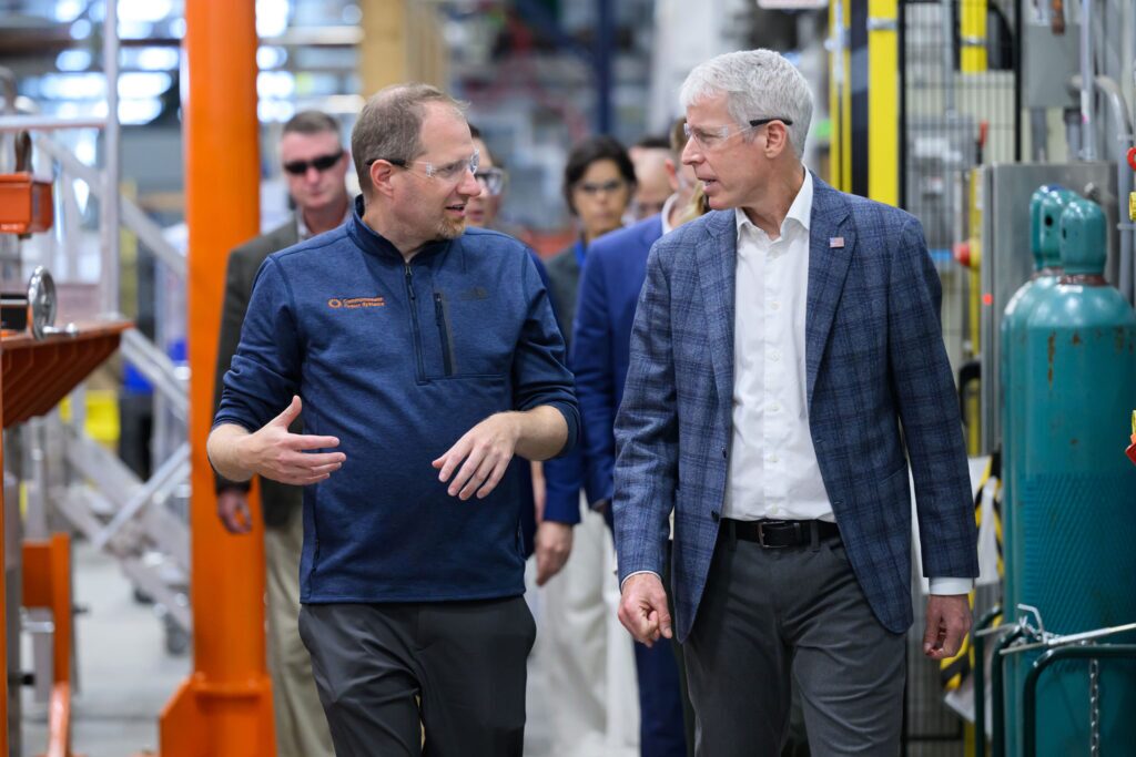 Two men wearing protective eyewear walk through a factory floor, orange metal structures on one side and green tanks of gas on the other.