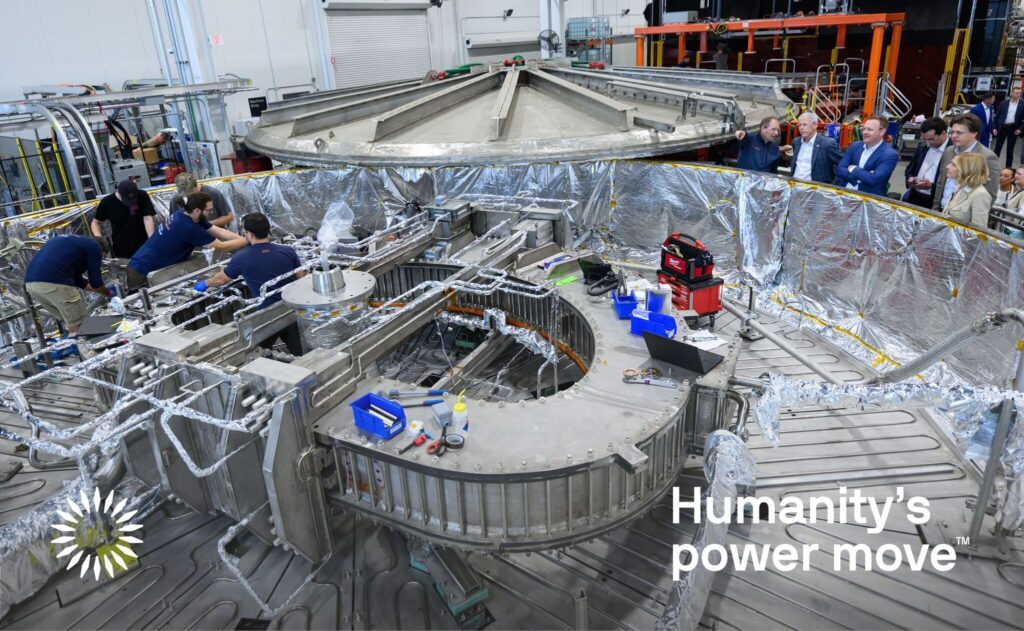A group of people discuss a large, wide silvery cylindrical test chamber that houses a D-shaped metal structure in its center. Five workers inside the chamber make adjustments. A number of tools and computers nearby support the operation