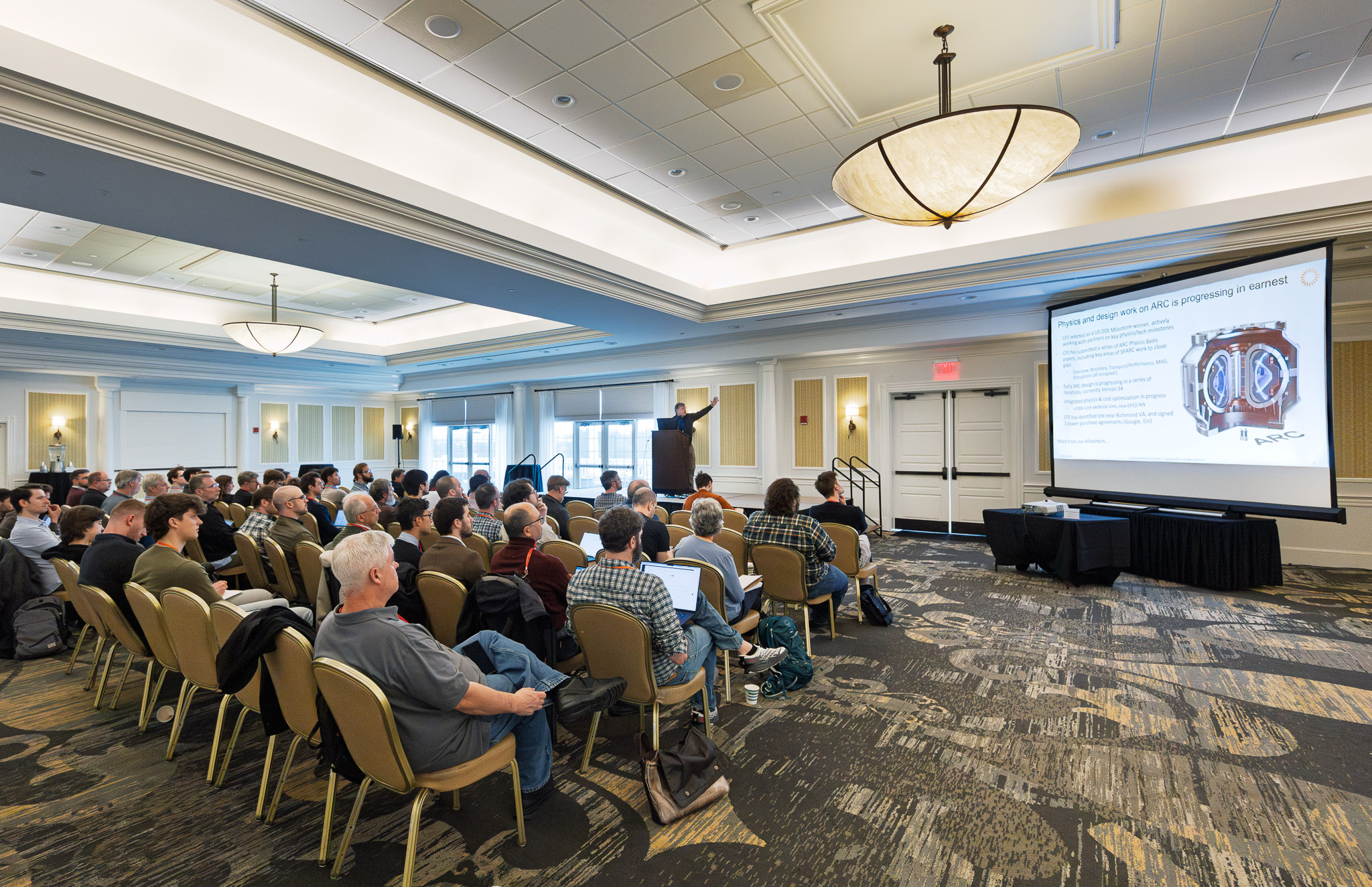 Conference room filled with attendees listening to a presenter at a podium, with a large projected slide on the screen behind them.