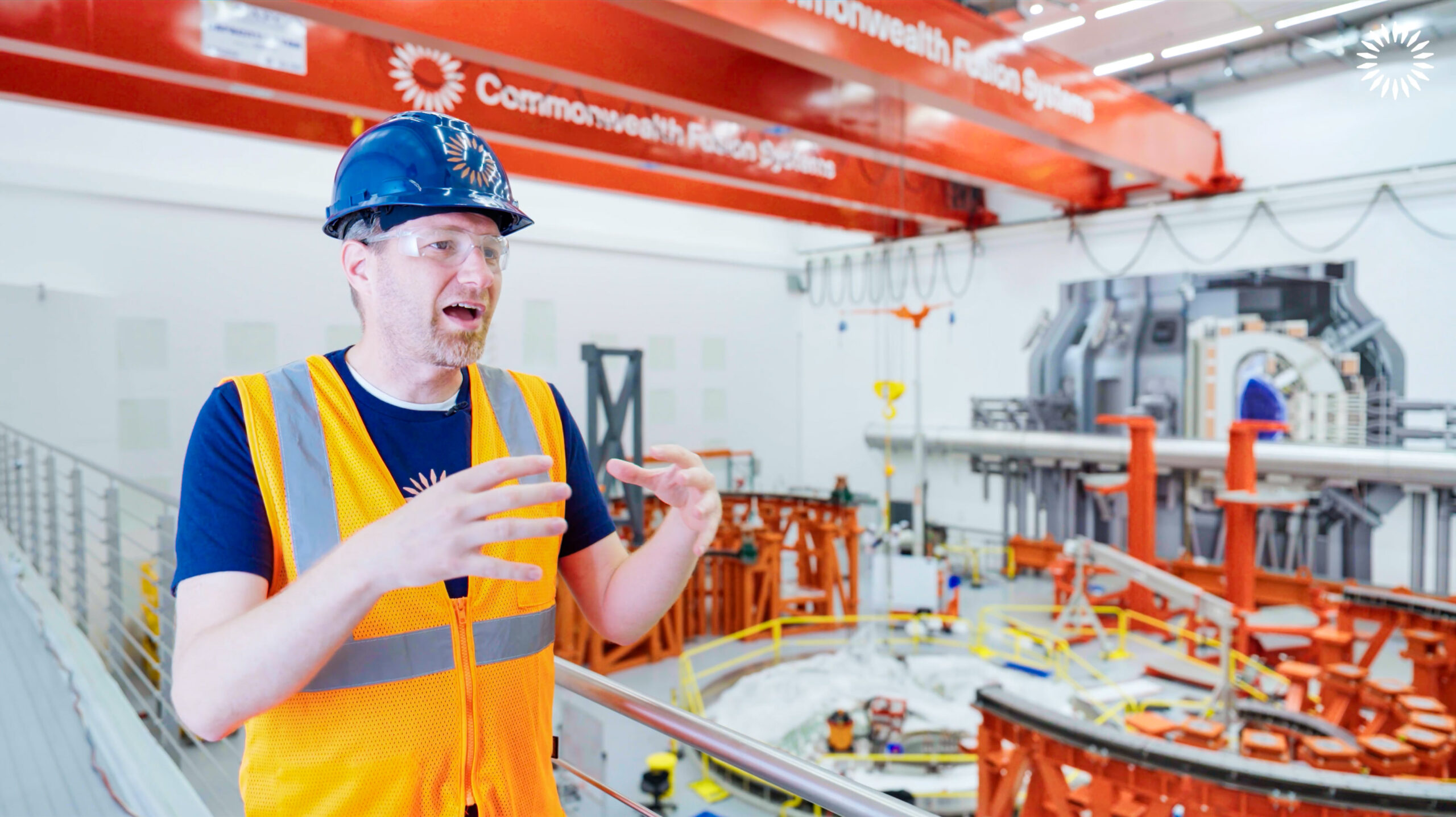 Commonwealth Fusion Systems CEO Bob Mumgaard, wearing an orange safety vest and blue hard hat, speaks in Tokamak Hall in front of the base of the SPARC tokamak.