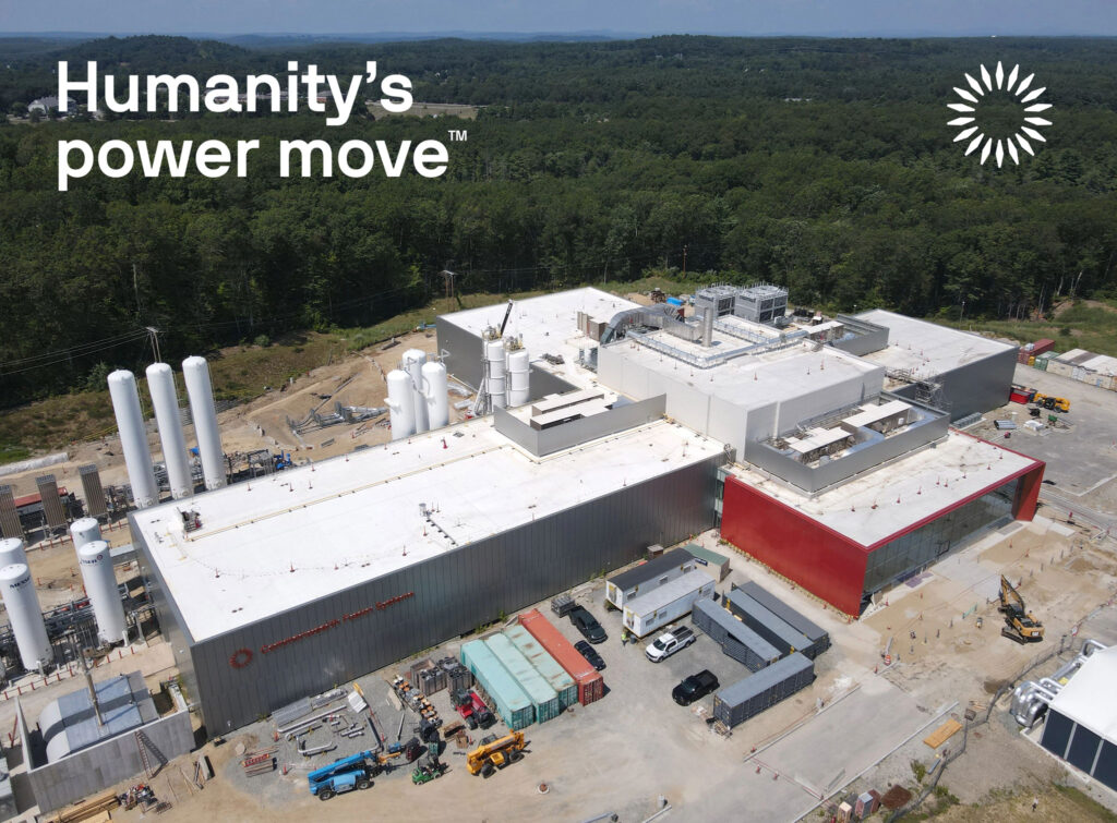 An aerial view of Commonwealth Fusion Systems' SPARC facility in Devens, Massachusetts, where the company is building a machine called a tokamak to demonstrate net fusion energy generation. The cross-shaped building has a taller central hall, is surrounded by tall tanks, and abuts a green forest.